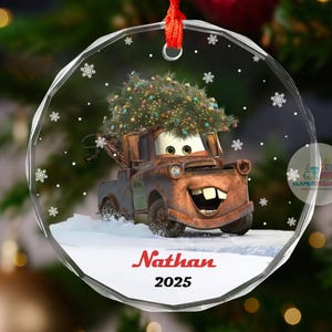 May include: A clear, round Christmas ornament featuring Mater from the movie Cars, with a Christmas tree on top. The ornament has snowflakes and the name "Nathan" and the year "2025" in red. It hangs from a red ribbon.
