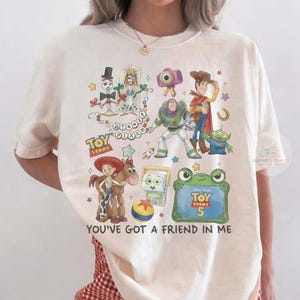 May include: A cream-colored t-shirt featuring a colorful graphic print of various Toy Story characters including Woody, Buzz Lightyear, Jessie, and Forky. The design includes the text Disney Pixar Toy Story and You've Got A Friend In Me.