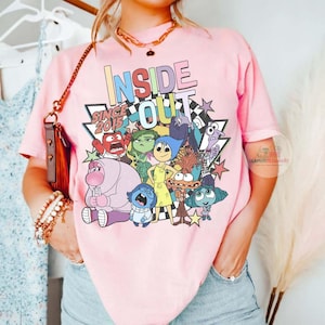 Pu&ograve; includere: T-shirt rosa chiaro con la scritta "INSIDE OUT" e la frase "SINCE 2015", oltre a una grafica dei personaggi del film d'animazione. Il design include una stella e un motivo a scacchi.