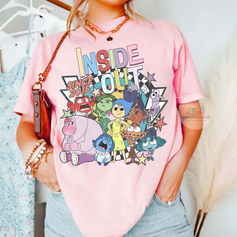 Pu&ograve; includere: T-shirt rosa chiaro con la scritta "INSIDE OUT" e la frase "SINCE 2015", oltre a una grafica dei personaggi del film d'animazione. Il design include una stella e un motivo a scacchi.