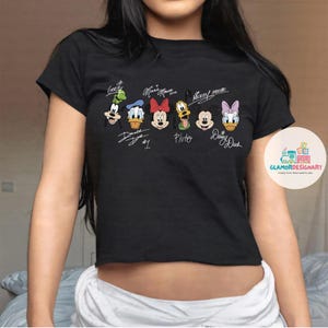 Puede incluir: Camiseta negra recortada con un gr&aacute;fico de Mickey Mouse y sus amigos, incluyendo a Donald Duck, Goofy, Pluto y Daisy Duck. Cada personaje tiene su firma debajo de su imagen.