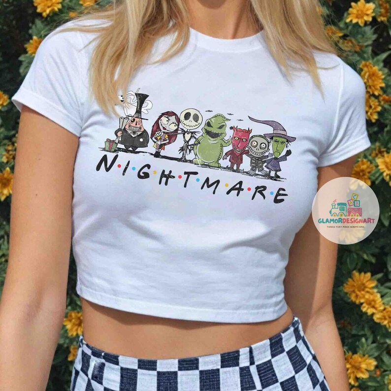 Może przedstawiać: Biały top z napisem "NIGHTMARE" czarnymi literami i postaciami z kresk&oacute;wek z filmu "Miasteczko Halloween". Projekt przedstawia postacie z filmu.