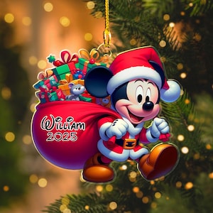 May include: A Christmas ornament featuring Mickey Mouse in a Santa Claus outfit, carrying a red sack filled with gifts. The ornament has the name "William" and the year "2025" printed on it. The background is a blurred image of a Christmas tree.