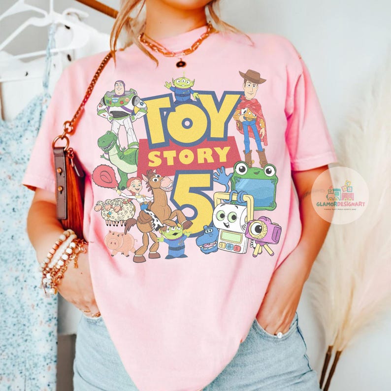 K&ouml;nnte beinhalten: Ein hellrosa T-Shirt mit einem farbenfrohen Grafikdruck der Toy Story 5-Figuren, darunter Woody, Buzz Lightyear, Jessie, Bullseye, Rex, Hamm und die Aliens. Das Design zeigt den fetten gelben und roten Schriftzug Toy Story 5.