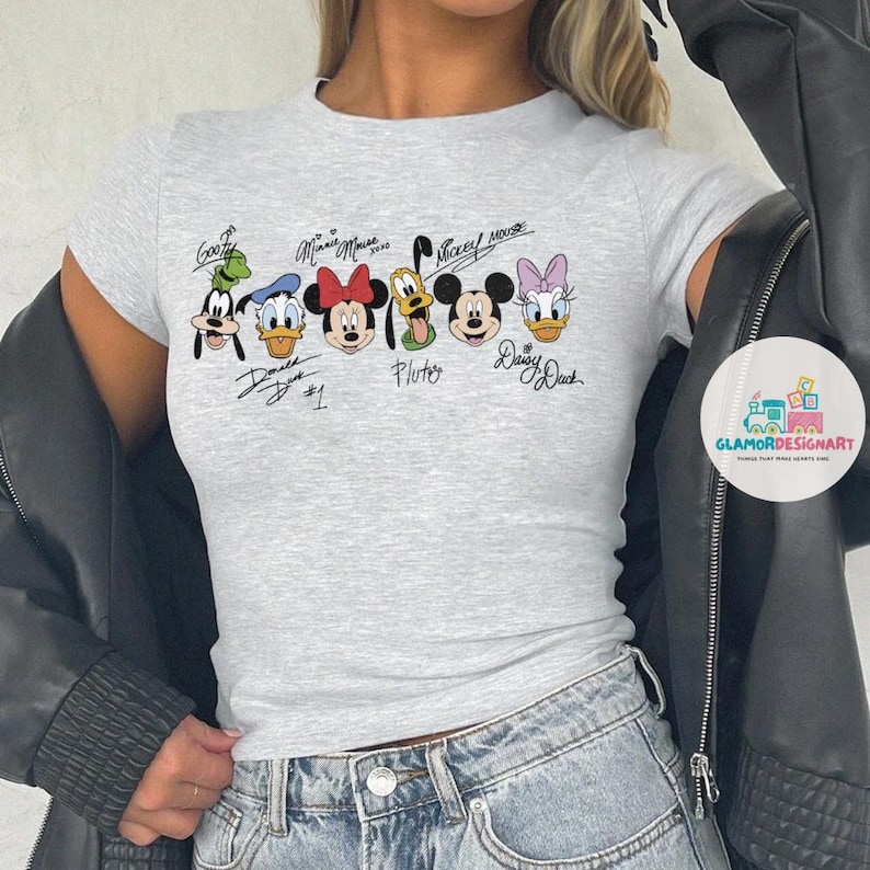 Puede incluir: Una camiseta gris con un estampado gr&aacute;fico de Mickey Mouse, Minnie Mouse, Donald Duck, Goofy, Pluto y Daisy Duck. Los personajes est&aacute;n en l&iacute;nea y tienen sus nombres escritos debajo.