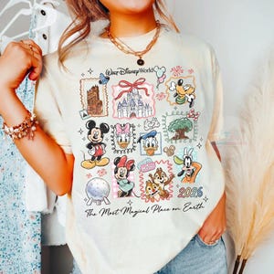 May include: Cream-colored t-shirt featuring various Disney characters and landmarks, including Mickey Mouse, Minnie Mouse, and Cinderella's castle. The shirt has the text "Walt Disney World" and "2026" with the phrase "The Most Magical Place on Earth".