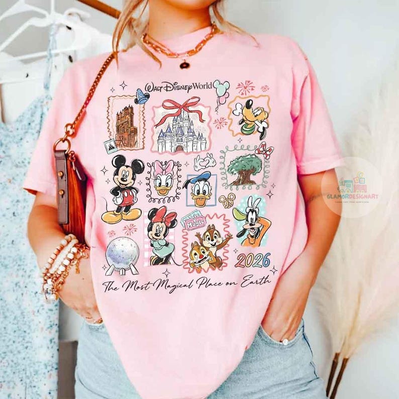 May include: Pink t-shirt featuring a colorful graphic of Disney characters and landmarks. The design includes Mickey Mouse, Minnie Mouse, Donald Duck, and Chip and Dale, with the text "Walt Disney World" and "2026". The shirt also has the text "The Most Magical Place on Earth".