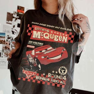 Może przedstawiać: Ciemnoszara koszulka z Lightning McQueen z filmu Disney Pixar Cars. Grafika zawiera tekst "Radiator Springs Saturday Night Special Race" i "Winner Takes All". Projekt jest w kolorach czerwonym, białym i czarnym.
