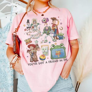 May include: A light pink t-shirt featuring a colorful graphic print of Toy Story characters including Woody, Buzz Lightyear, Jessie, Bullseye, Forky, and an alien. The design includes the text Toy Story, Disney Pixar, and You've Got A Friend In Me.