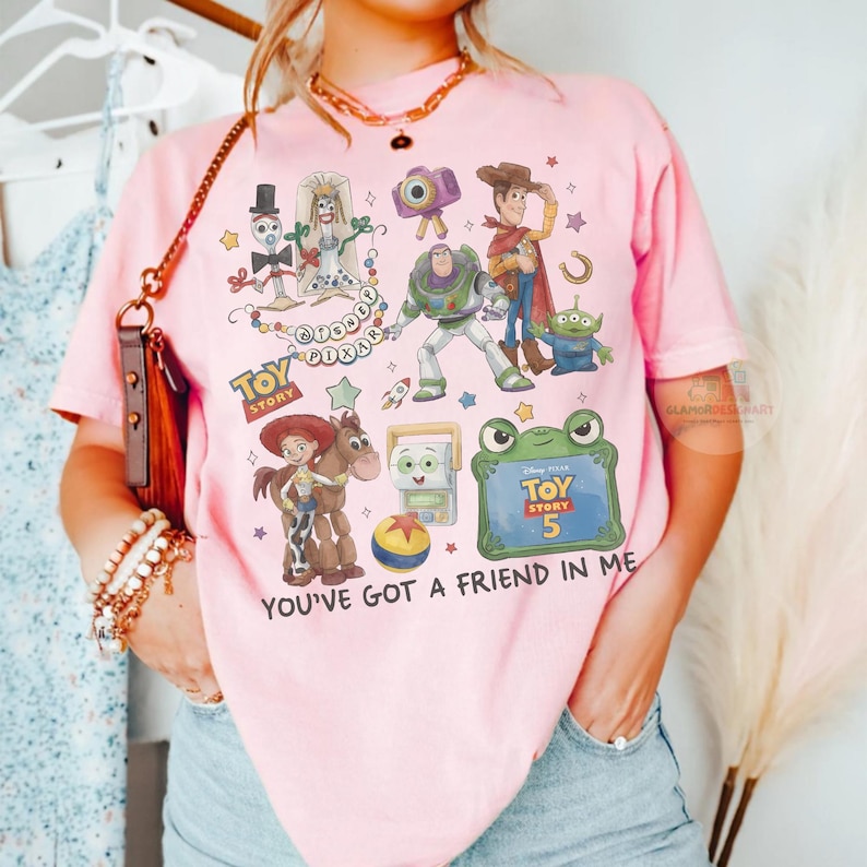 May include: A light pink t-shirt featuring a colorful graphic print of Toy Story characters including Woody, Buzz Lightyear, Jessie, Bullseye, Forky, and an alien. The design includes the text Toy Story, Disney Pixar, and You've Got A Friend In Me.