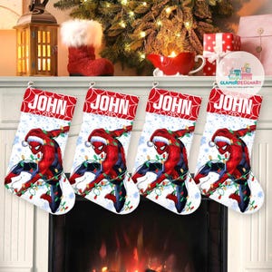 May include: Four Christmas stockings with the name "JOHN" in red letters. Each stocking features a graphic of Spider-Man wearing a Santa hat and wrapped in Christmas lights. The stockings are white with a red top and are hanging above a fireplace.