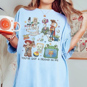 May include: A light blue t-shirt featuring a graphic print of Toy Story characters including Woody, Buzz Lightyear, Jessie, Bullseye, and Forky. The design includes the text Toy Story, Disney Pixar, and You've Got A Friend In Me, along with a Toy Story 5 logo.