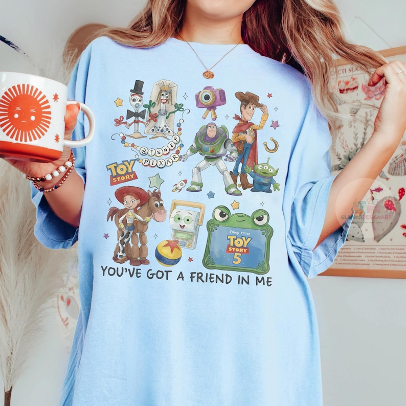 May include: A light blue t-shirt featuring a graphic print of Toy Story characters including Woody, Buzz Lightyear, Jessie, Bullseye, and Forky. The design includes the text Toy Story, Disney Pixar, and You've Got A Friend In Me, along with a Toy Story 5 logo.