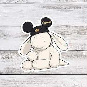 May include: A sticker of a cartoon bunny wearing a black hat with the name "Thomas" in gold lettering. The bunny is white with long ears and black Mickey Mouse-style ears. The sticker is on a white wooden background.