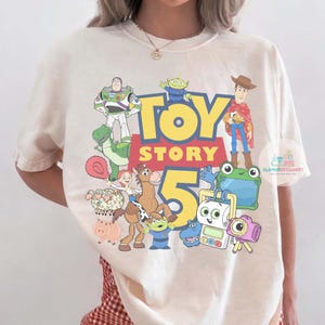K&ouml;nnte beinhalten: Ein cremefarbenes Kurzarm-T-Shirt mit einem bunten Grafikdruck der Toy Story 5-Figuren, darunter Buzz Lightyear, Woody, Jessie, Bullseye, Rex und verschiedene andere animierte Charaktere mit dem gro&szlig;en gelb-roten Toy Story 5-Logo.