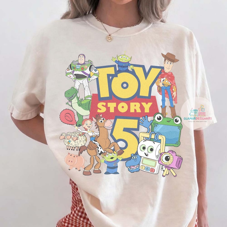 K&ouml;nnte beinhalten: Ein cremefarbenes Kurzarm-T-Shirt mit einem bunten Grafikdruck der Toy Story 5-Figuren, darunter Buzz Lightyear, Woody, Jessie, Bullseye, Rex und verschiedene andere animierte Charaktere mit dem gro&szlig;en gelb-roten Toy Story 5-Logo.
