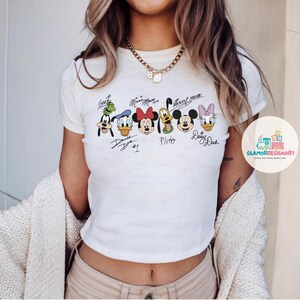 Puede incluir: Camiseta blanca recortada con un gr&aacute;fico de Mickey Mouse, Minnie Mouse, Donald Duck, Goofy y Pluto. Los personajes est&aacute;n en l&iacute;nea y tienen sus nombres escritos debajo.
