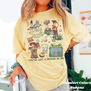 May include: A pale yellow t-shirt featuring a colorful graphic print of Toy Story characters including Woody, Buzz Lightyear, Jessie, Bullseye, and Forky. The design includes the text Toy Story, Disney Pixar, Toy Story 5, and You've Got A Friend In Me.