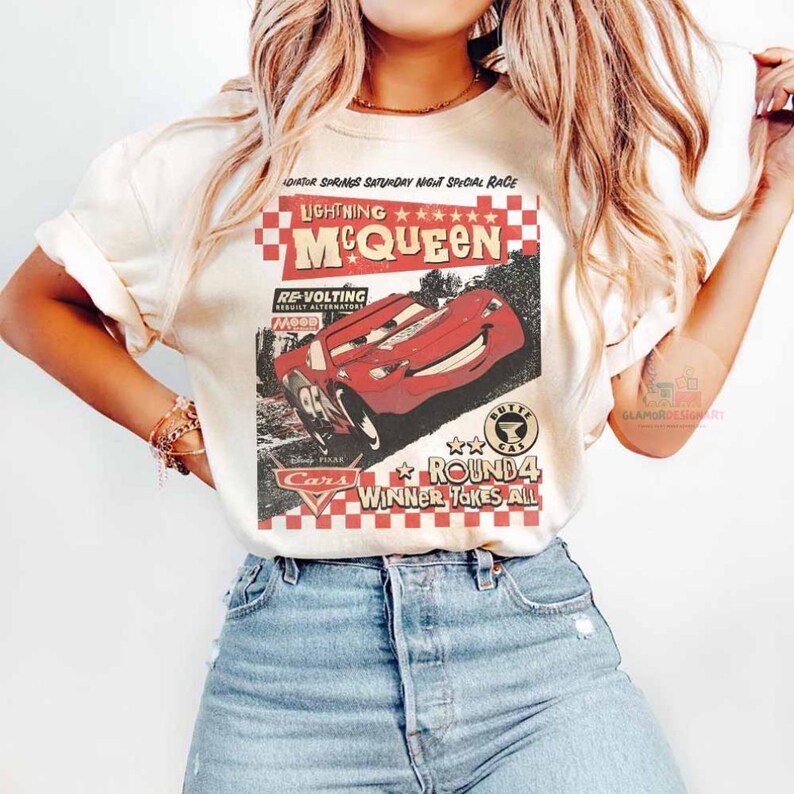 Może przedstawiać: Kremowa koszulka z grafiką Zygzaka McQueena z filmu Auta. Projekt zawiera tekst "Lightning McQueen" i "Round 4 Winner Takes All" w stylu vintage. Koszulka jest połączona z niebieskimi dżinsami.