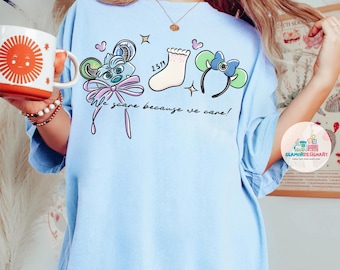 May include: Light blue t-shirt with a whimsical design featuring a mouse, a sock, and a headband with a bow. The shirt has the text "We scare because we care!" printed on it. A white mug with a sun design is held in the left hand.