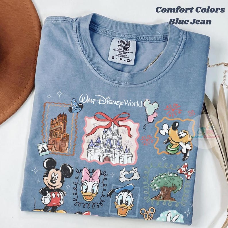 May include: Blue jean Comfort Colors t-shirt featuring a colorful graphic of Walt Disney World characters and landmarks. The design includes Mickey Mouse, Daisy Duck, Donald Duck, and a castle. The shirt is a size small.