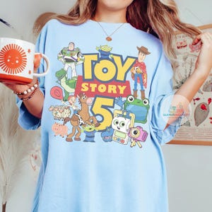 K&ouml;nnte beinhalten: Ein hellblaues T-Shirt mit einem farbenfrohen Grafikdruck von Toy Story-Figuren wie Woody, Buzz Lightyear, Jessie und Bullseye, wobei der Text Toy Story 5 in gro&szlig;en gelben und roten Buchstaben auf der Vorderseite des Kleidungsst&uuml;cks zentriert ist.