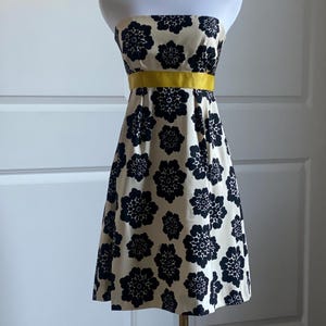 May include: A strapless dress on a mannequin. The dress features a cream-colored background with a repeating pattern of navy blue floral designs. A bright yellow band accents the waist. The dress is knee-length.