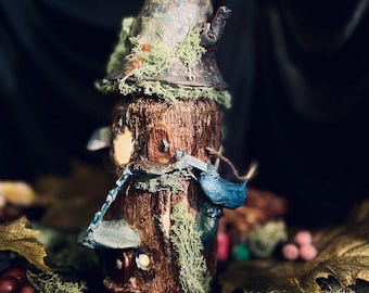 May include: A miniature treehouse figurine with a moss-covered roof and a blue and white birdhouse attached. The treehouse is made of wood and has a rustic, whimsical style.