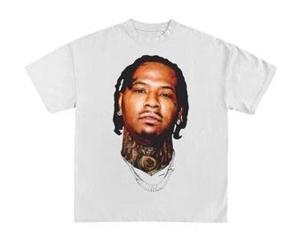 May include: White t-shirt featuring a detailed portrait of a man with tattoos on his neck and a silver chain. The image is a close-up of the man's face, showcasing his hairstyle and facial features. The shirt is plain and simple.