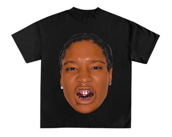 May include: A black short-sleeved t-shirt featuring a large, centered graphic print of a person's face. The person has a septum piercing, ear studs, and a lip piercing, and is wearing dark-colored dental grills on their teeth.