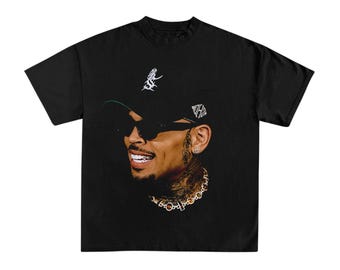 May include: A black short-sleeve t-shirt featuring a graphic print of a smiling person wearing a black baseball cap with white text, dark sunglasses, and a gold chain necklace. The person has visible neck and facial tattoos and a small earring.