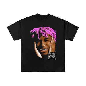 Puede incluir: Una camiseta negra de manga corta con un estampado gráfico de una persona con rastas de color rosa brillante, las manos enmarcando la cara y grillz plateados en los dientes. Debajo del rostro hay un emblema tribal blanco circular.