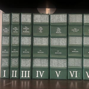 May include: A collection of seven hardcover books with green covers, featuring titles like "The Sorcerer's Stone" and "The Deathly Hallows." Each book has a white diamond pattern and the author's name, J.K. Rowling, printed on the spine. Roman numerals I through VII are at the bottom.