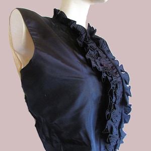 Puede incluir: Un top sin mangas de color azul marino con un cuello alto con volantes. El top está hecho de una tela lisa, posiblemente satinada. El cuello con volantes añade un elemento decorativo a la prenda.