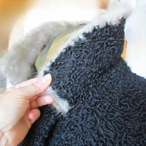 May include: A black curly fur coat with a grey fur collar. The collar is turned up and the fur is soft and fluffy.