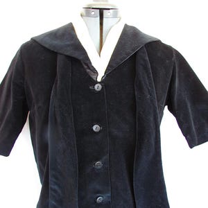 Könnte beinhalten: Schwarze Samtjacke mit breitem Kragen und drei Knöpfen vorne. Die Jacke hat kurze Ärmel und eine weiße Unterschicht, die am Ausschnitt sichtbar ist. Das Kleidungsstück wird auf einer Schaufensterpuppe präsentiert.