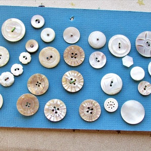 May include: A collection of 20 vintage mother of pearl buttons in various shapes and sizes. The buttons are arranged on a blue background.
