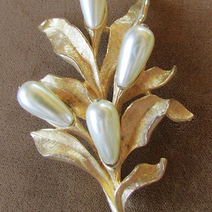 May include: A vintage gold-tone brooch in the shape of a stylized leaf arrangement. The brooch features four pearlescent, teardrop-shaped faux pearls. The leaves have detailed veining, and the overall design is elegant.