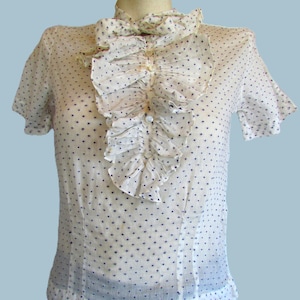 Könnte beinhalten: Eine weiße Bluse mit kurzen Ärmeln und einem hochgeschlossenen Kragen mit Rüschen. Die Bluse ist mit kleinen marineblauen Punkten verziert. Die Vorderseite hat eine vertikale Rüschendetail und wird mit kleinen Knöpfen geschlossen. Das Kleidungsstück ist im Vintage-Stil.