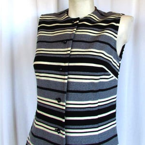 May include: Sleeveless vest with horizontal stripes in black, white, and gray. The vest has a button-up front and a tailored fit. The garment is displayed on a mannequin, showcasing its design and style. A classic piece for layering.