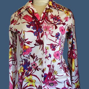 May include: White button-down shirt with a vibrant floral pattern. The long-sleeved shirt features a colorful design of pink, red, yellow, and blue flowers and foliage. The shirt has a classic collar and button closure.