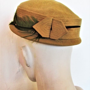 Peut inclure: Un chapeau en feutre beige avec une garniture de plumes vertes et un nœud décoratif sur le côté.