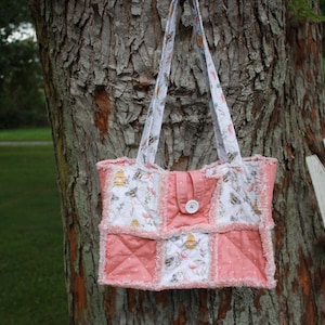 May include: A patchwork tote bag featuring alternating squares of solid coral pink fabric and white fabric printed with bees and honeycombs. The bag has frayed seams, two matching shoulder straps, and a central tab closure secured with a white button.