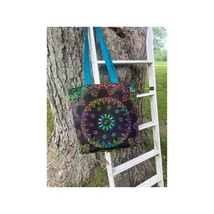 May include: Black tote bag with a colorful, circular design. The design features a rainbow of colors, including green, blue, purple, and yellow. The bag has bright blue handles and is hanging on a tree.