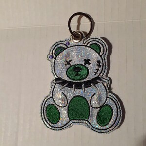 May include: A silver glitter bear keychain with green eyes, a black studded collar, and a stitched X over each eye.