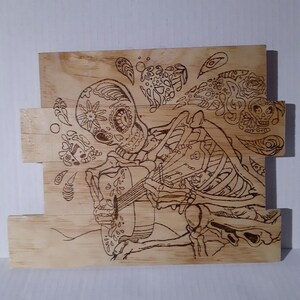 May include: A wood burned design of a skeleton playing a guitar with a sugar skull face. The design is surrounded by floral and paisley patterns.