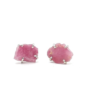 May include: A pair of pink tourmaline gemstone stud earrings. Each earring features a raw, irregular-shaped pink gemstone held in place by a silver-coloured metal setting. The earrings are set against a white background.