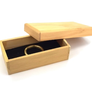 Può includere: Una scatola di legno con un coperchio, aperta per rivelare un anello d'oro all'interno. La scatola è in legno chiaro e ha un rivestimento in velluto nero.