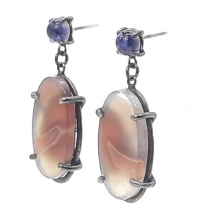 Peut inclure: Une paire de boucles d'oreilles pendantes en métal argenté. Chaque boucle d'oreille présente une pierre ovale de couleur pêche clair avec un motif unique, surmontée d'une petite pierre carrée bleu foncé. Les boucles d'oreilles sont pour oreilles percées.