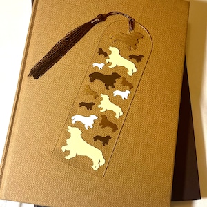 May include: A gold bookmark with a tassel, featuring a silhouette of a dachshund dog in various sizes and colors. The bookmark is on a brown book.